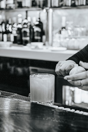 A busy bar scene with a focus on a cocktail being prepared. A hand squeezes a citrus fruit over a glass filled with ice. The background features shelves stocked with bottles and glassware, conveying an upscale ambiance.