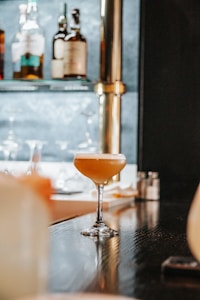 A cocktail glass containing a light brown beverage is placed on a polished dark bar counter. In the background, there are blurred bottles on a shelf, suggesting a bar setting. The atmosphere appears sophisticated and well-lit, with reflections visible on the bar's surface.