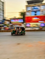 A green and yellow auto rickshaw is moving quickly along a city street. The background is blurred, indicating motion, with buildings featuring bright advertisements and signs. The road and surrounding environment have a busy, urban feel.