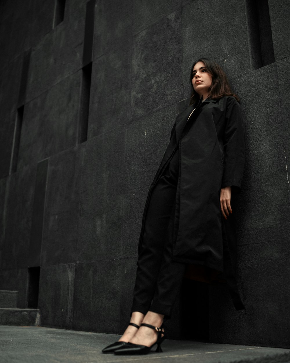 a woman leaning against a wall wearing a black coat