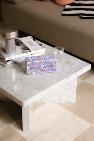 A marble coffee table topped with a stack of books, a silver cylindrical lamp, a colorful patterned object, and a glass of water. Part of a sofa with a striped fabric visible in the background.