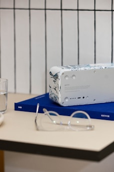 A white speaker with a marbled design on its sides sits on a blue book titled 'BOULDER' on a beige table. Nearby, a pair of transparent glasses is placed next to a clear glass of water. The background features a white tiled wall with thin black lines.