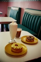 A cozy cafe setting with retro-style teal seating and a tiled wall. On a white table, there are two donuts on yellow plates, one topped with icing and the other with chocolate and sprinkles. There is also a glass of milkshake with whipped cream and a cup of coffee.