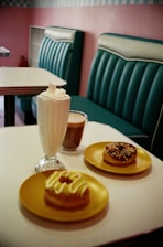 A cozy cafe setting with retro-style teal seating and a tiled wall. On a white table, there are two donuts on yellow plates, one topped with icing and the other with chocolate and sprinkles. There is also a glass of milkshake with whipped cream and a cup of coffee.