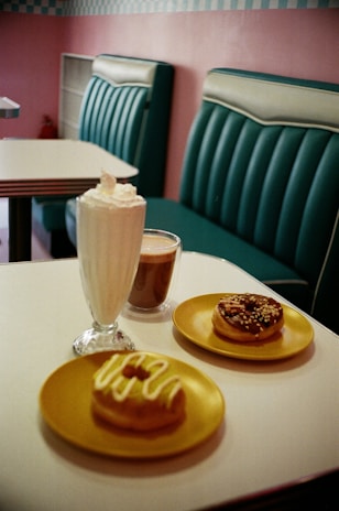 A cozy cafe setting with retro-style teal seating and a tiled wall. On a white table, there are two donuts on yellow plates, one topped with icing and the other with chocolate and sprinkles. There is also a glass of milkshake with whipped cream and a cup of coffee.