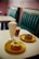 A cozy cafe setting with retro-style teal seating and a tiled wall. On a white table, there are two donuts on yellow plates, one topped with icing and the other with chocolate and sprinkles. There is also a glass of milkshake with whipped cream and a cup of coffee.