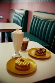A cozy cafe setting with retro-style teal seating and a tiled wall. On a white table, there are two donuts on yellow plates, one topped with icing and the other with chocolate and sprinkles. There is also a glass of milkshake with whipped cream and a cup of coffee.