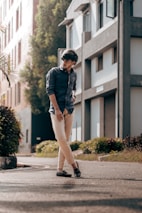 A young man is standing on a sunlit urban street. He is wearing a dark blue shirt and beige pants, coupled with black loafers. Behind him are modern buildings with large windows and greenery along the sidewalk, creating a casual yet stylish urban setting.