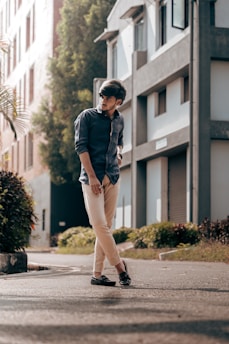 A young man is standing on a sunlit urban street. He is wearing a dark blue shirt and beige pants, coupled with black loafers. Behind him are modern buildings with large windows and greenery along the sidewalk, creating a casual yet stylish urban setting.
