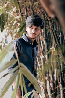 A young man with styled hair is standing amidst tall bamboo plants, wearing a dark blue shirt. The light filters through the leaves, casting natural patterns on him.