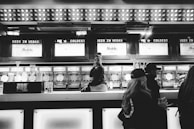 A bar setting with a person sitting on the counter, surrounded by frozen drink dispensers labeled with various flavors. The bar has bright lights above and a sign reading 'Beer in Vegas'. Several people are nearby, engaged in conversation.
