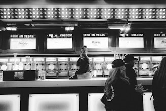 A bar setting with a person sitting on the counter, surrounded by frozen drink dispensers labeled with various flavors. The bar has bright lights above and a sign reading 'Beer in Vegas'. Several people are nearby, engaged in conversation.