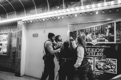 A group of people stand near a wall under bright marquee lights, one of whom appears to be a security officer holding another person's arm. A large poster for Hugo's Cellar restaurant is visible in the background.