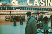 An older man with a gray beard and wearing a suit and hat stands in front of a brightly lit casino. The background features a large illuminated sign reading 'CASINO' and 'SLOTS' among other words, with numerous lights outlining the entrance. Several other people are visible in the background, engaged in various activities, some walking, some standing, and one looking at something on display.