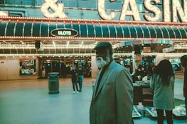 An older man with a gray beard and wearing a suit and hat stands in front of a brightly lit casino. The background features a large illuminated sign reading 'CASINO' and 'SLOTS' among other words, with numerous lights outlining the entrance. Several other people are visible in the background, engaged in various activities, some walking, some standing, and one looking at something on display.