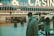 An older man with a gray beard and wearing a suit and hat stands in front of a brightly lit casino. The background features a large illuminated sign reading 'CASINO' and 'SLOTS' among other words, with numerous lights outlining the entrance. Several other people are visible in the background, engaged in various activities, some walking, some standing, and one looking at something on display.