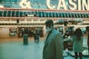 An older man with a gray beard and wearing a suit and hat stands in front of a brightly lit casino. The background features a large illuminated sign reading 'CASINO' and 'SLOTS' among other words, with numerous lights outlining the entrance. Several other people are visible in the background, engaged in various activities, some walking, some standing, and one looking at something on display.