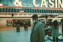 An older man with a gray beard and wearing a suit and hat stands in front of a brightly lit casino. The background features a large illuminated sign reading 'CASINO' and 'SLOTS' among other words, with numerous lights outlining the entrance. Several other people are visible in the background, engaged in various activities, some walking, some standing, and one looking at something on display.