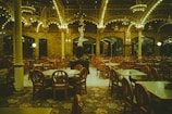 An empty restaurant featuring vintage-style round wooden chairs and tables arranged neatly under a high ceiling adorned with ornate ironwork and glowing chandeliers. The walls are made of brick, contributing to a warm and classic ambiance.