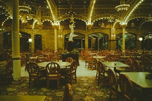 An empty restaurant featuring vintage-style round wooden chairs and tables arranged neatly under a high ceiling adorned with ornate ironwork and glowing chandeliers. The walls are made of brick, contributing to a warm and classic ambiance.