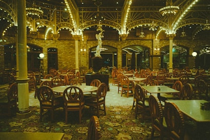 An empty restaurant featuring vintage-style round wooden chairs and tables arranged neatly under a high ceiling adorned with ornate ironwork and glowing chandeliers. The walls are made of brick, contributing to a warm and classic ambiance.