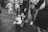 A black and white photo capturing a busy street scene with several people walking. A child is sitting in a stroller with a neutral expression, while an adult pushes the stroller. Other individuals are engaged with their phones or in conversation. The scene is urban and dynamic, suggesting a casual day in a city.