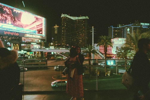 A bustling cityscape at night with illuminated billboards and neon lights. Several people are visible in the foreground, adding to the lively urban atmosphere. Prominent signage includes various restaurants and hotels surrounded by palm trees.