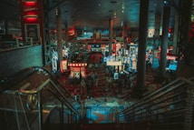 A bustling casino floor filled with groups of slot machines and people walking around. The atmosphere is vibrant with neon lights and illuminated signs. The interior has a ceiling supported by large pillars and there are stairs leading down to the gaming area.
