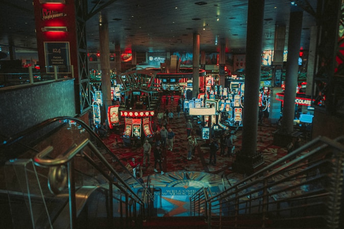 A bustling casino floor filled with groups of slot machines and people walking around. The atmosphere is vibrant with neon lights and illuminated signs. The interior has a ceiling supported by large pillars and there are stairs leading down to the gaming area.
