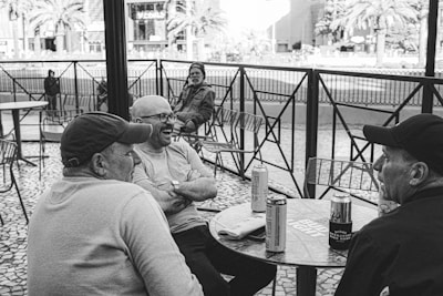 Three men are sitting at an outdoor cafe table, engaged in conversation and laughter. They are accompanied by cans of beverage on the table. Chairs and a tiled pavement create a casual, relaxed atmosphere. In the background, a man sits alone with palm trees and city buildings visible.