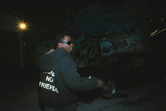 A sleek, dark-themed photo of a model wearing an oversized hoodie with a glowing neon silhouette print, standing against a minimalist urban backdrop at night.