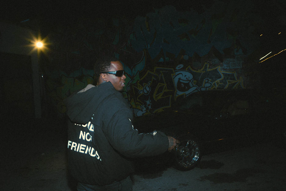 Urban streetwear model wearing black hoodie with neon accents, standing against a graffiti wall at dusk.