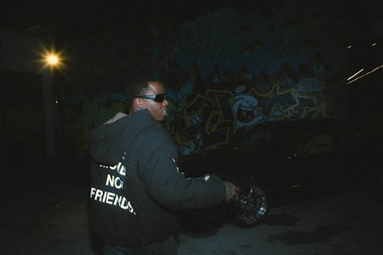 A person wearing dark sunglasses and a black hoodie with white text stands in a dimly lit urban setting. The background features vibrant graffiti art and a bright light source shines from the left side, creating stark contrasts and shadows.