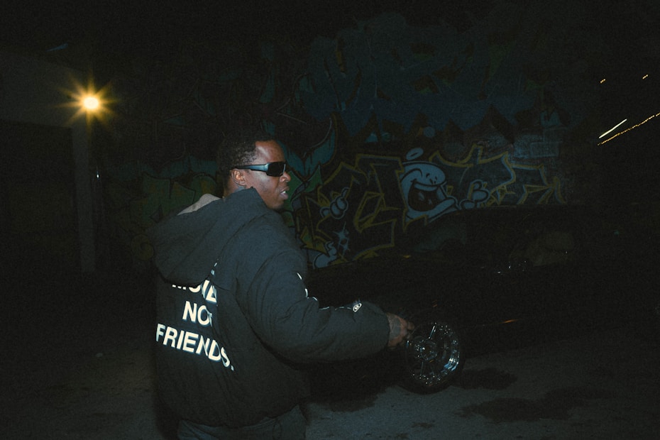 A person wearing dark sunglasses and a black hoodie with white text stands in a dimly lit urban setting. The background features vibrant graffiti art and a bright light source shines from the left side, creating stark contrasts and shadows.