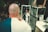 A bald man wearing a stylish two-toned suit is standing in front of a mirror, adjusting his attire and taking a selfie. He stands in a well-lit, modern room with various objects, including a display case with jewelry, a figure, a laptop, and posters or advertisements.