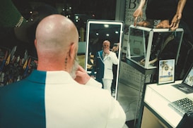 A bald man wearing a stylish two-toned suit is standing in front of a mirror, adjusting his attire and taking a selfie. He stands in a well-lit, modern room with various objects, including a display case with jewelry, a figure, a laptop, and posters or advertisements.