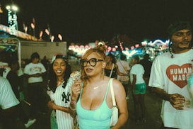 A group of people are enjoying themselves at a nighttime event or fair. Bright lights from amusement rides illuminate the scene, while a woman in focus is eating ice cream and wearing large glasses. Others around her seem relaxed and happy, adding to the festive atmosphere.