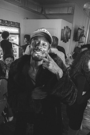 A group of people gather in a dimly lit room with a man standing prominently in the foreground, smiling and making a peace sign gesture. Behind him, other people are casually interacting near a beverage cooler, and some are seated. The setting appears relaxed and informal.
