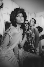 A black and white photograph of three individuals in a room with minimalist decor. The person in the foreground wears a fur headpiece and a dress, striking a confident pose. The other two individuals stand closely behind, each with distinct expressions giving off an artistic and edgy vibe. The background shows some simple furniture and a window.
