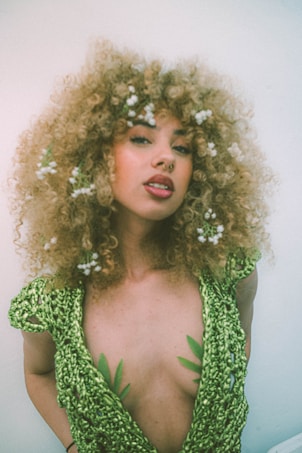 A person with curly hair adorned with small white flowers is wearing a green textured outfit with a deep neckline. The attire features leaf-like patterns, creating a natural and organic theme.