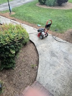 A red and black pressure washer is placed on a concrete pathway next to a green lawn. There are garden plants and small shrubs bordering the path, with a garden hose connected to the washer. A broom is lying on the grass nearby, and the surrounding area appears to be well-maintained.