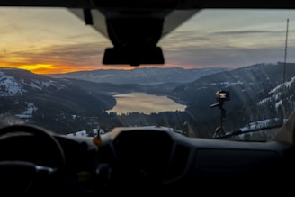 A scenic view from the interior of a vehicle showcases a mountainous landscape at sunset. Snow-covered mountains surround a serene lake, reflecting the soft hues of the evening sky. The dashboard and a mounted camera are faintly visible in the foreground.