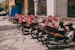 A row of motorbikes parked along a sidewalk, each equipped with a large red delivery box advertising a pizza company. The boxes feature images of pizza and promotional text. The scene is urban with a building and a few trees in the background.