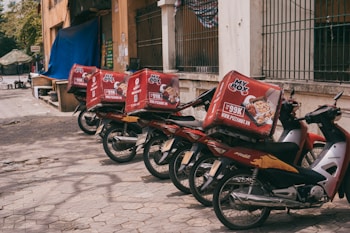 A row of motorbikes parked along a sidewalk, each equipped with a large red delivery box advertising a pizza company. The boxes feature images of pizza and promotional text. The scene is urban with a building and a few trees in the background.