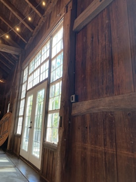 A rustic interior featuring wooden walls and large windows allowing natural light to stream in. The space appears cozy and warm, with soft lighting from small overhead bulbs. A white-framed door is centered within the window panels, providing a view of the greenery outside.