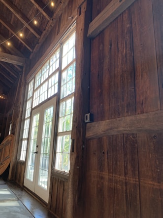 A rustic interior featuring wooden walls and large windows allowing natural light to stream in. The space appears cozy and warm, with soft lighting from small overhead bulbs. A white-framed door is centered within the window panels, providing a view of the greenery outside.