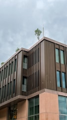 A modern building with a sleek design featuring brown and beige tones and large windows. Trees are growing on the rooftop, adding a touch of greenery against the cloudy sky.