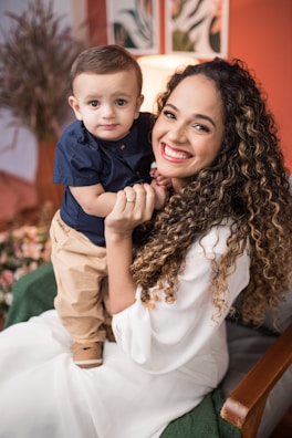 A woman with curly hair is smiling brightly while holding a baby dressed in a blue shirt and beige pants. They are both sitting on a chair in a warmly decorated room with art pieces and plants in the background.