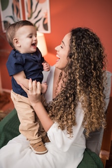 A woman with long, curly hair, wearing a white outfit, is holding a smiling baby boy dressed in a navy blue shirt and beige pants. They are both smiling and looking at each other in a warmly lit room with a reddish-brown wall and decorative paintings in the background.