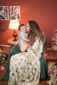 A woman with curly hair embraces a young child while sitting on a chair in a cozy, warmly lit room with a rustic, natural decor theme. The woman is wearing a long, patterned dress in green and white. Beside them is a table with a lit lamp, and floral arrangements decorate the surroundings.